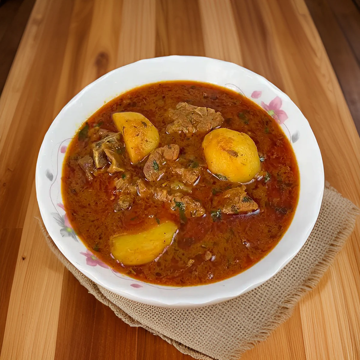 Aloo gosht in a white flowered bowl: meat and potatoes in a thin red gravy.