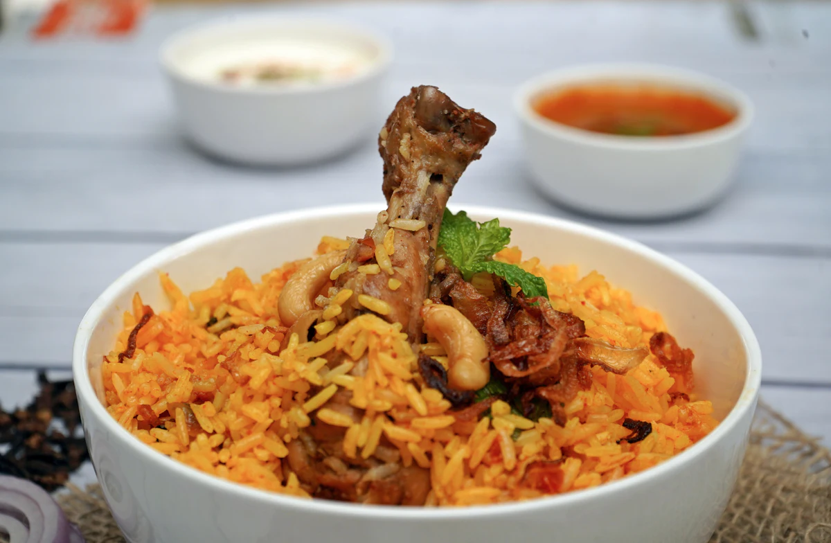 A white bowl of orange chicken biryani with a drumstick and fried cashews on top, raita behind it.