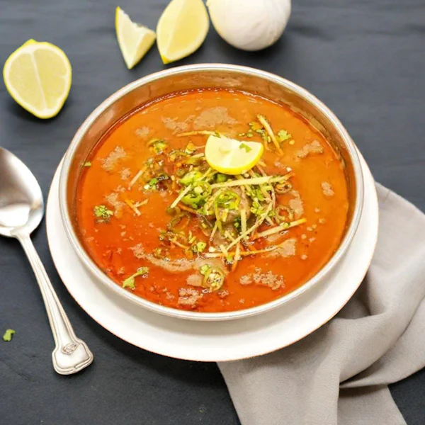 A bowl of orange haleem with fried onions, ginger and a lemon slice, a spoon beside it.