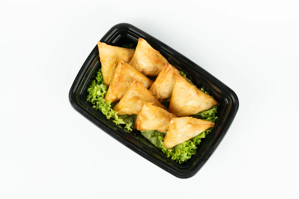 Six golden samosas on lettuce in a black tray on a white table.
